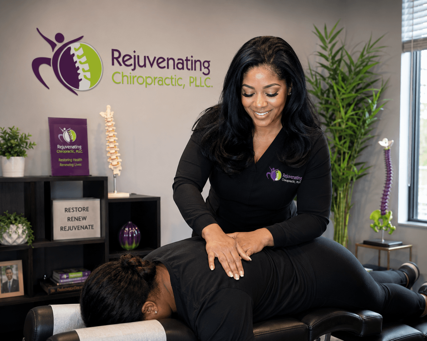 Chiropractor smiling while adjusting a patient's back in a clinic with plants and a logo on the wall.