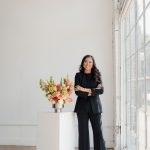 Woman in black suit standing next to a flower arrangement in a bright room with large windows