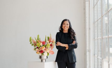 Woman in black suit standing next to a flower arrangement in a bright room with large windows