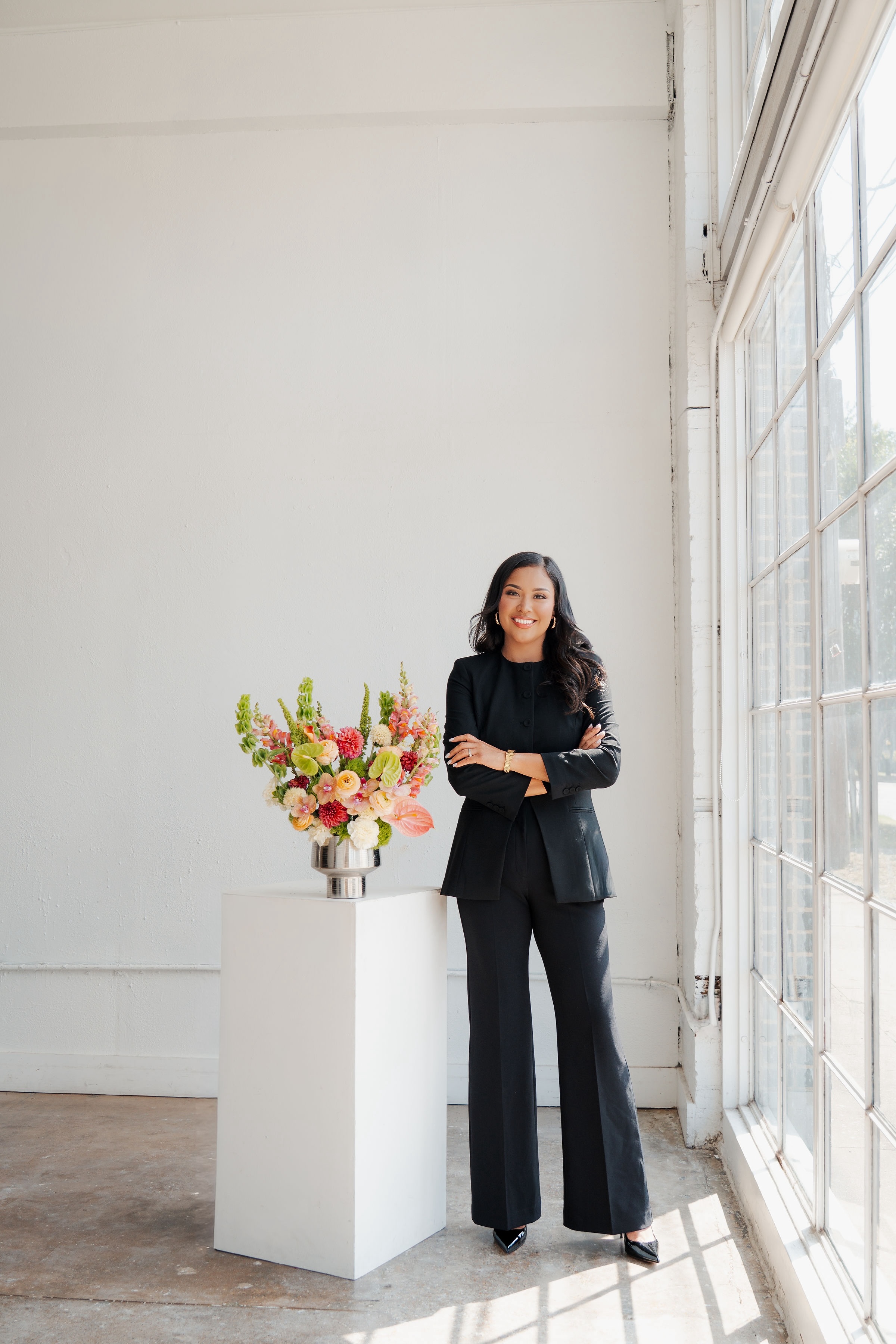 Woman in black suit standing next to a flower arrangement in a bright room with large windows