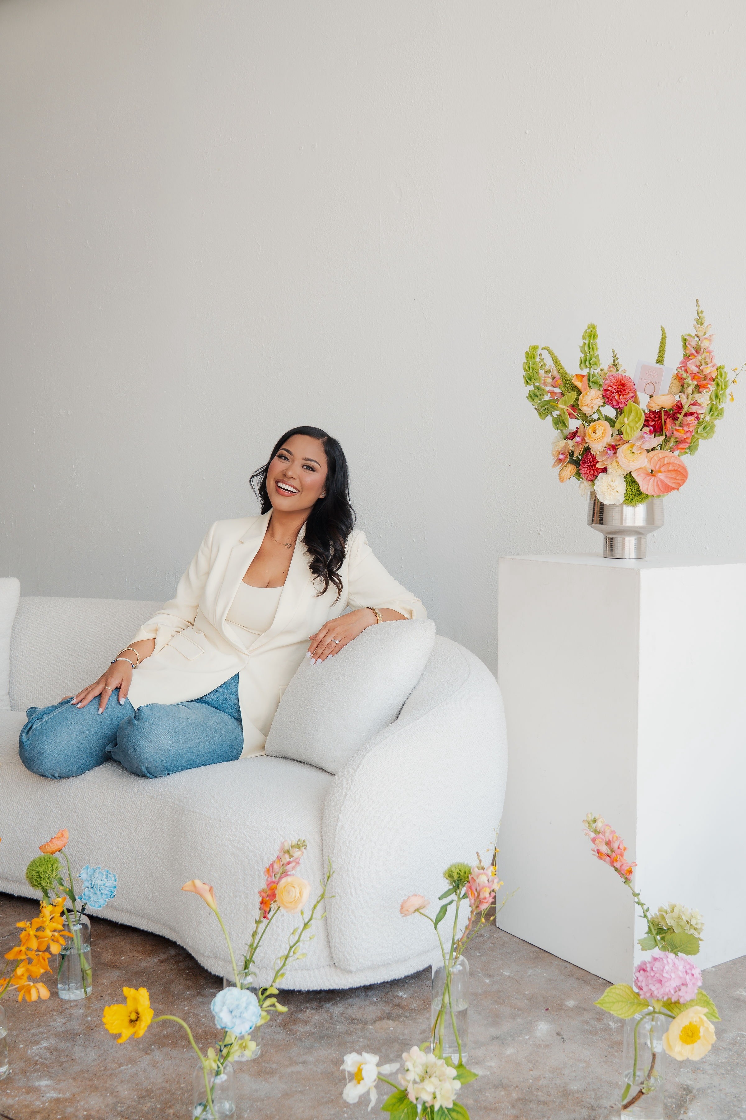 Woman sitting on a white sofa with a large flower arrangement on a pedestal beside her, smiling, in a bright room.