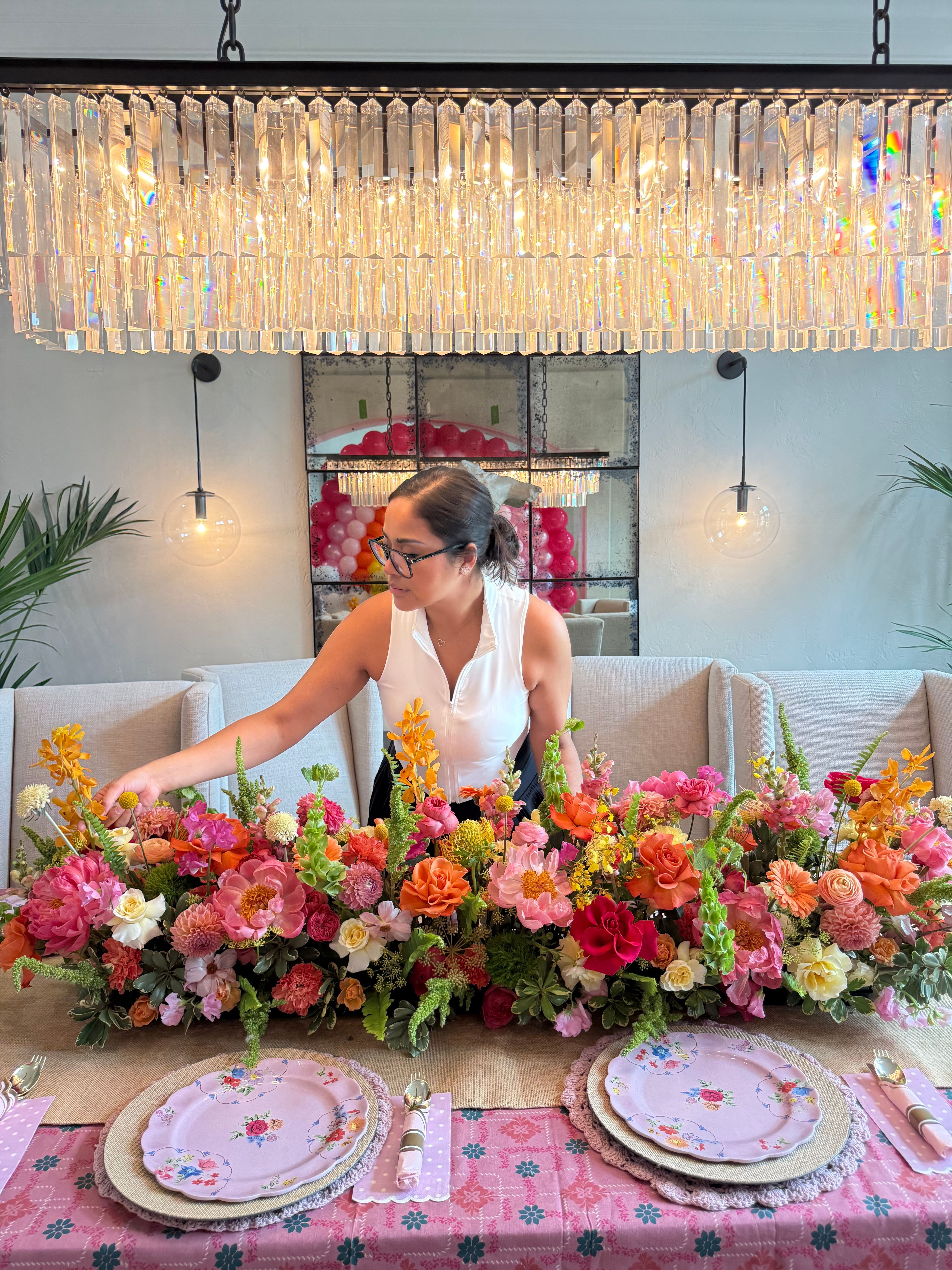 Woman arranging a large colorful flower centerpiece on a table with pink plates and napkins.