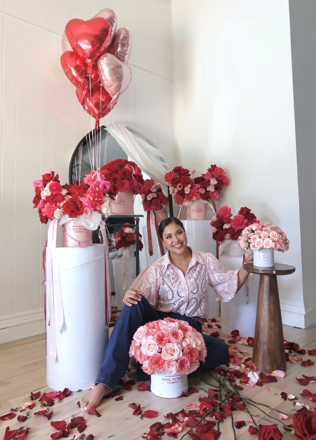Woman sitting on floor surrounded by pink and red flowers, with heart-shaped balloons above, in a decorated room.