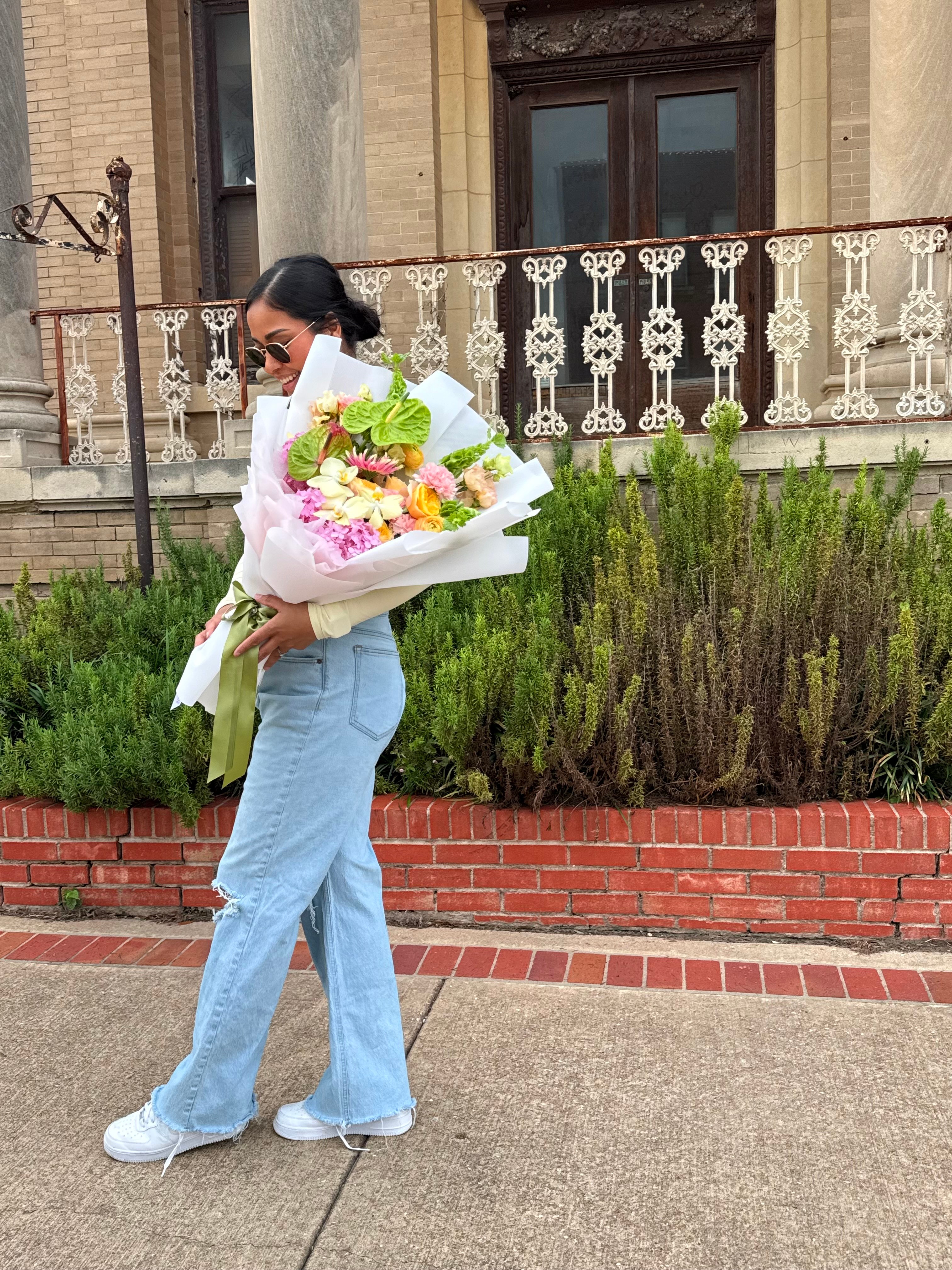Person holding a large bouquet of flowers standing outdoors in front of a building with a garden and brick border.