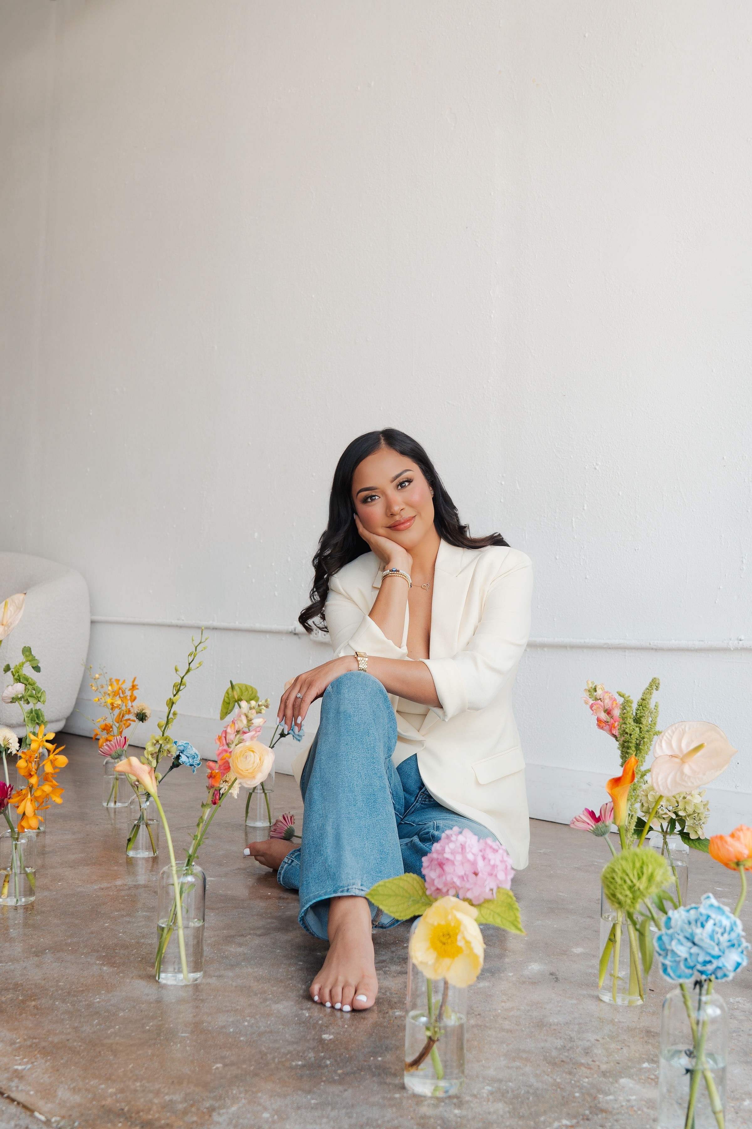 Woman sitting on floor surrounded by colorful flower vases, wearing white blazer and jeans, smiling, with a plain white wall background.