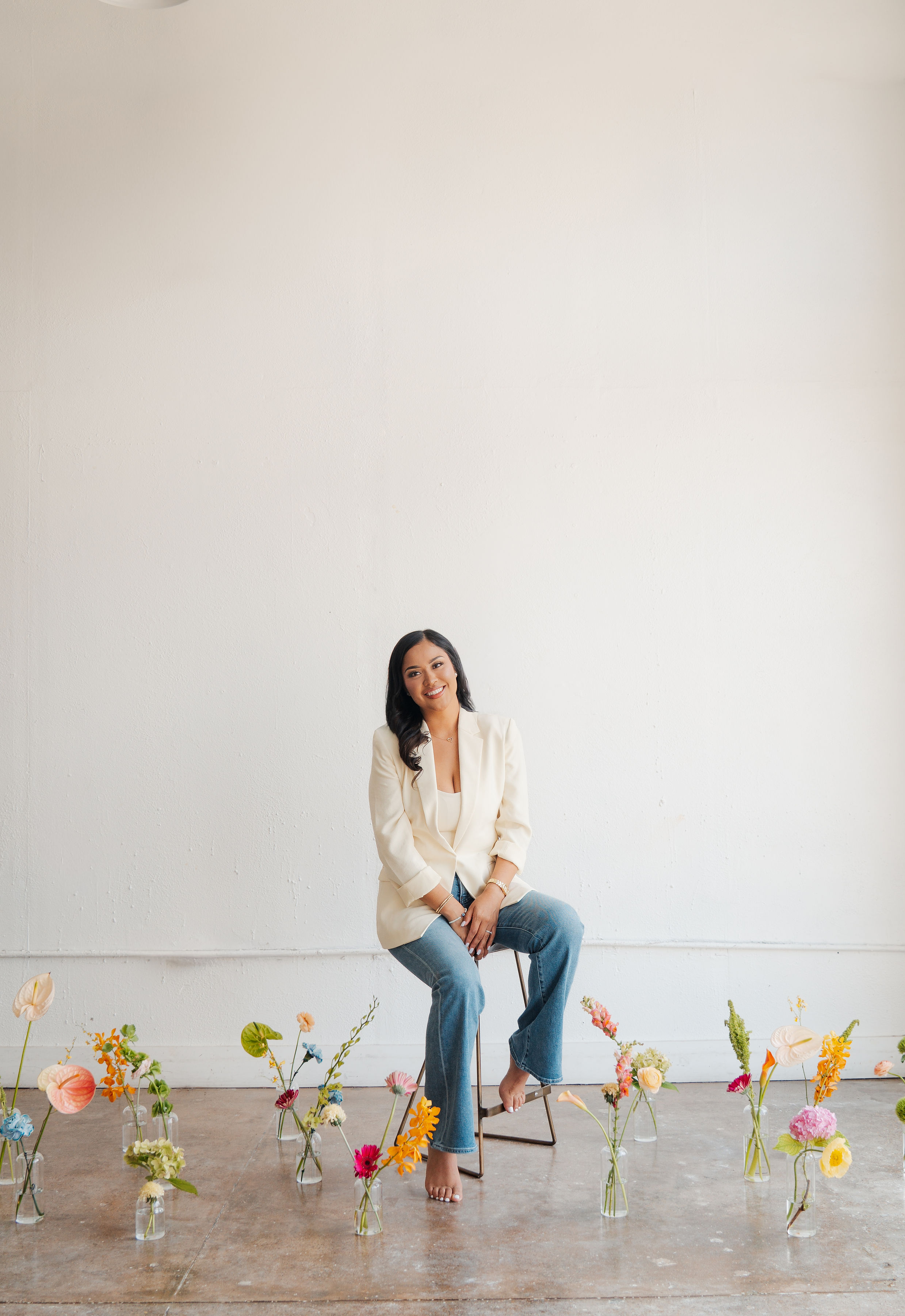 Woman sitting on a stool surrounded by colorful flowers in glass vases, against a plain white wall.