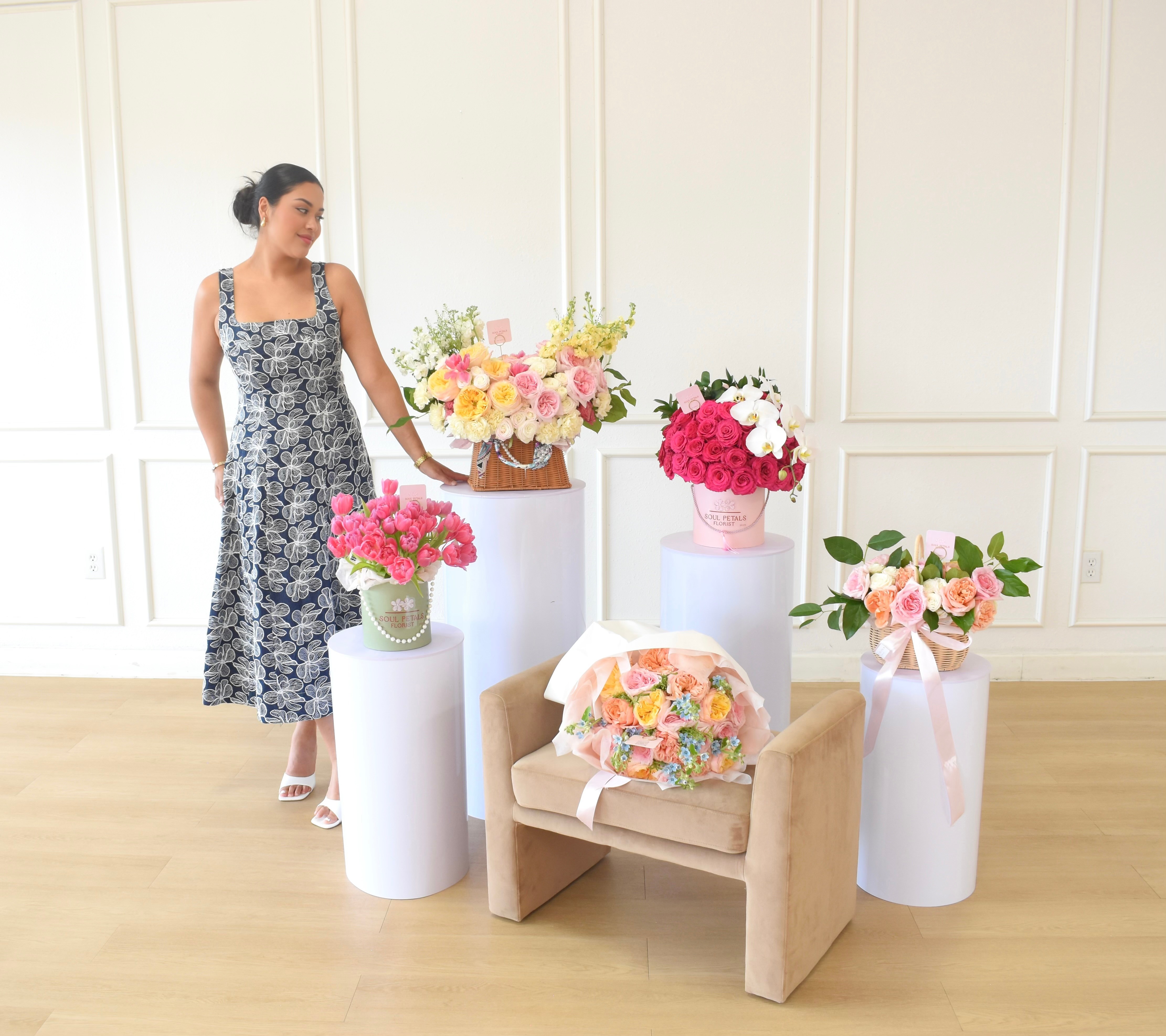 Woman in a floral dress standing next to flower arrangements on white pedestals in a bright room.