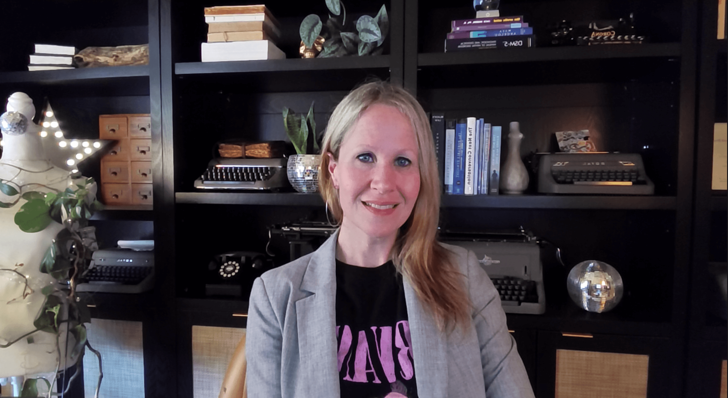 Woman with blonde hair smiling, sitting in front of a black bookshelf filled with books and decorative items.