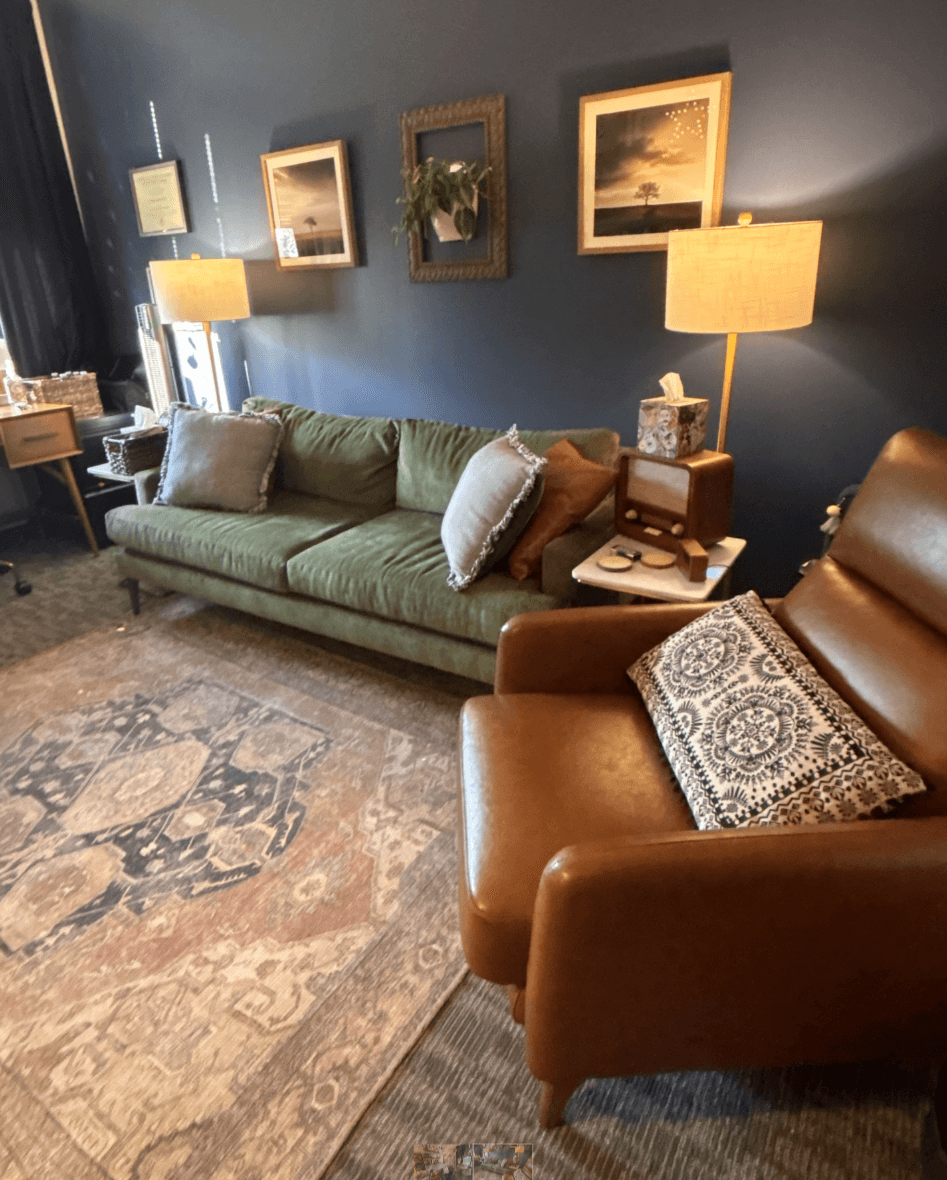 Living room with a green sofa, brown armchair, side table, lamps, framed pictures, and a patterned rug.