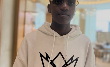 Person wearing sunglasses and a white hoodie with a crown logo and text, indoors with a blurred background.