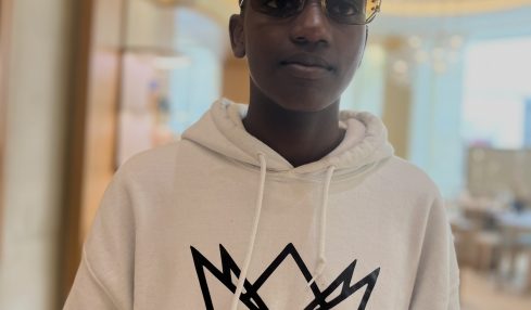 Person wearing sunglasses and a white hoodie with a crown logo and text, indoors with a blurred background.