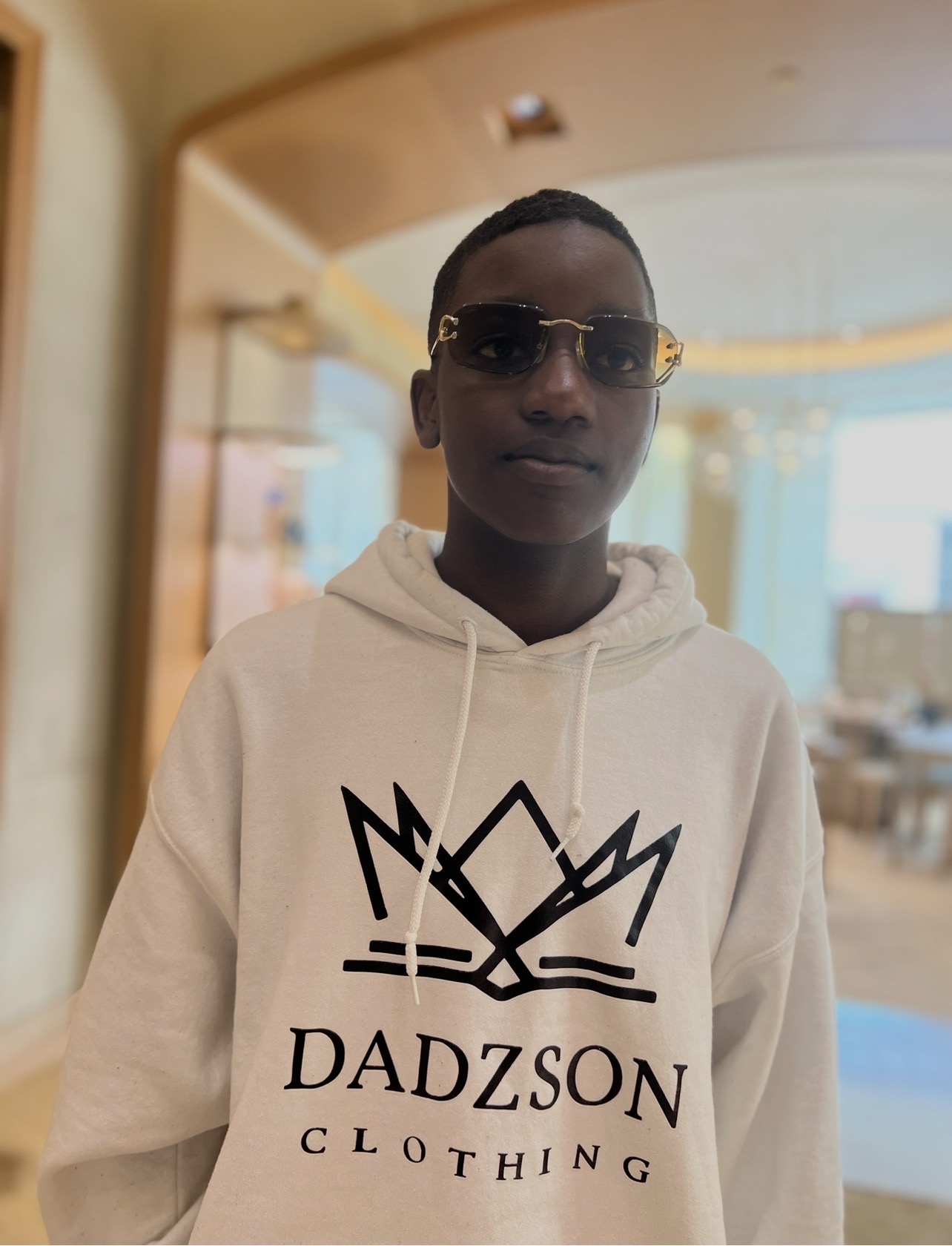 Person wearing sunglasses and a white hoodie with a crown logo and text, indoors with a blurred background.