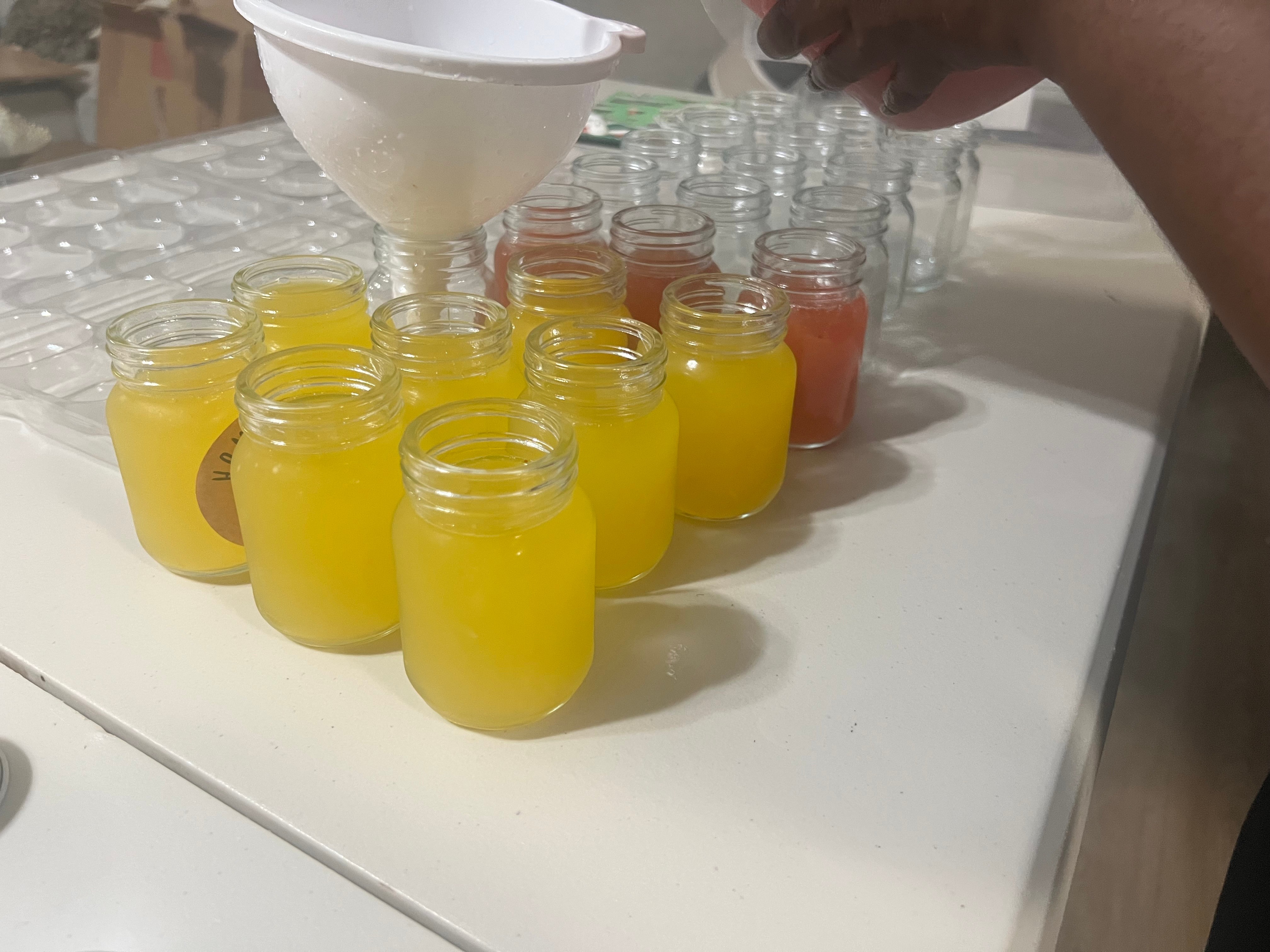 Person pouring yellow liquid into small glass jars on a white surface, with other jars in the background.