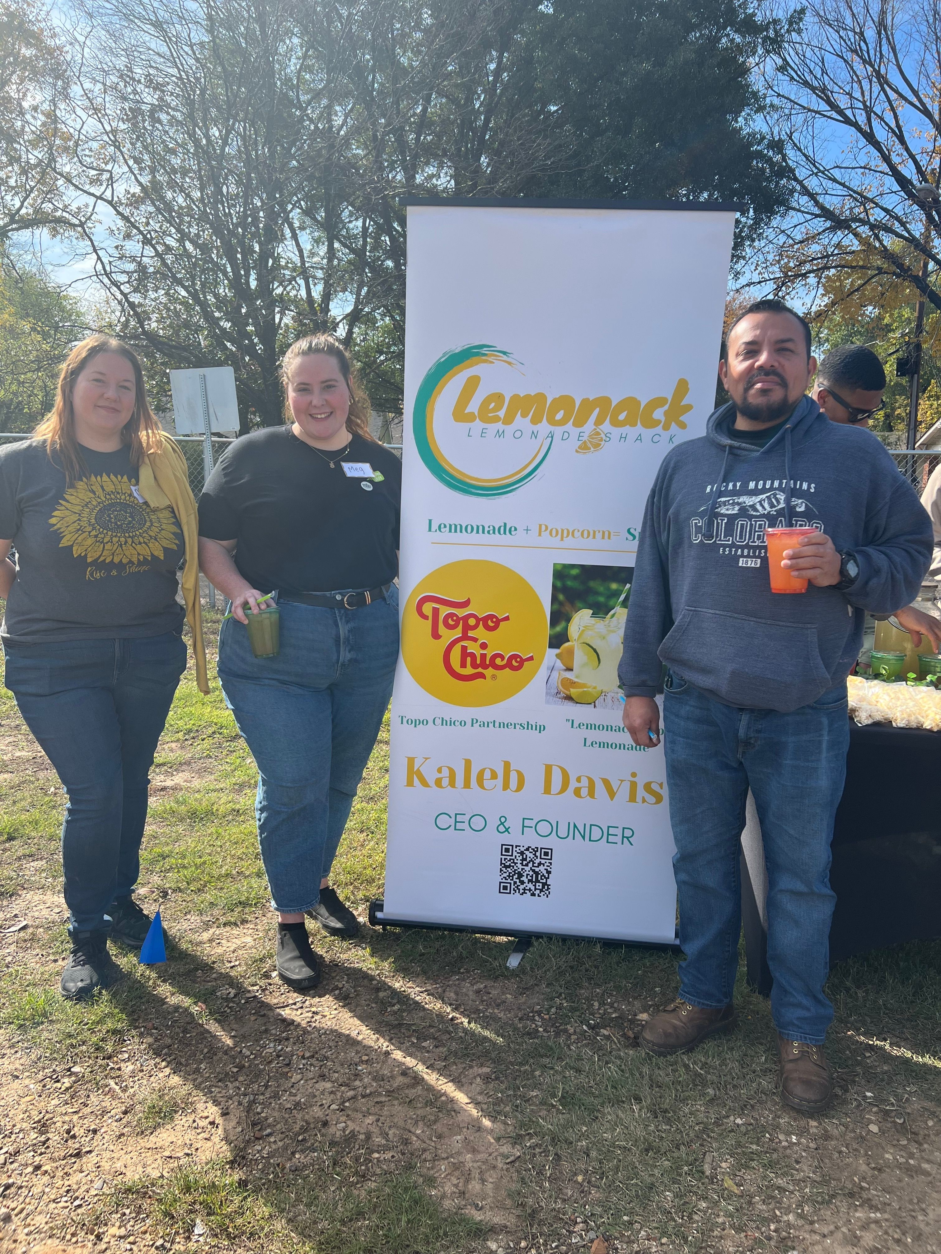 Three people standing outdoors near a signboard, trees in background, sunlight, casual clothing, holding drinks.