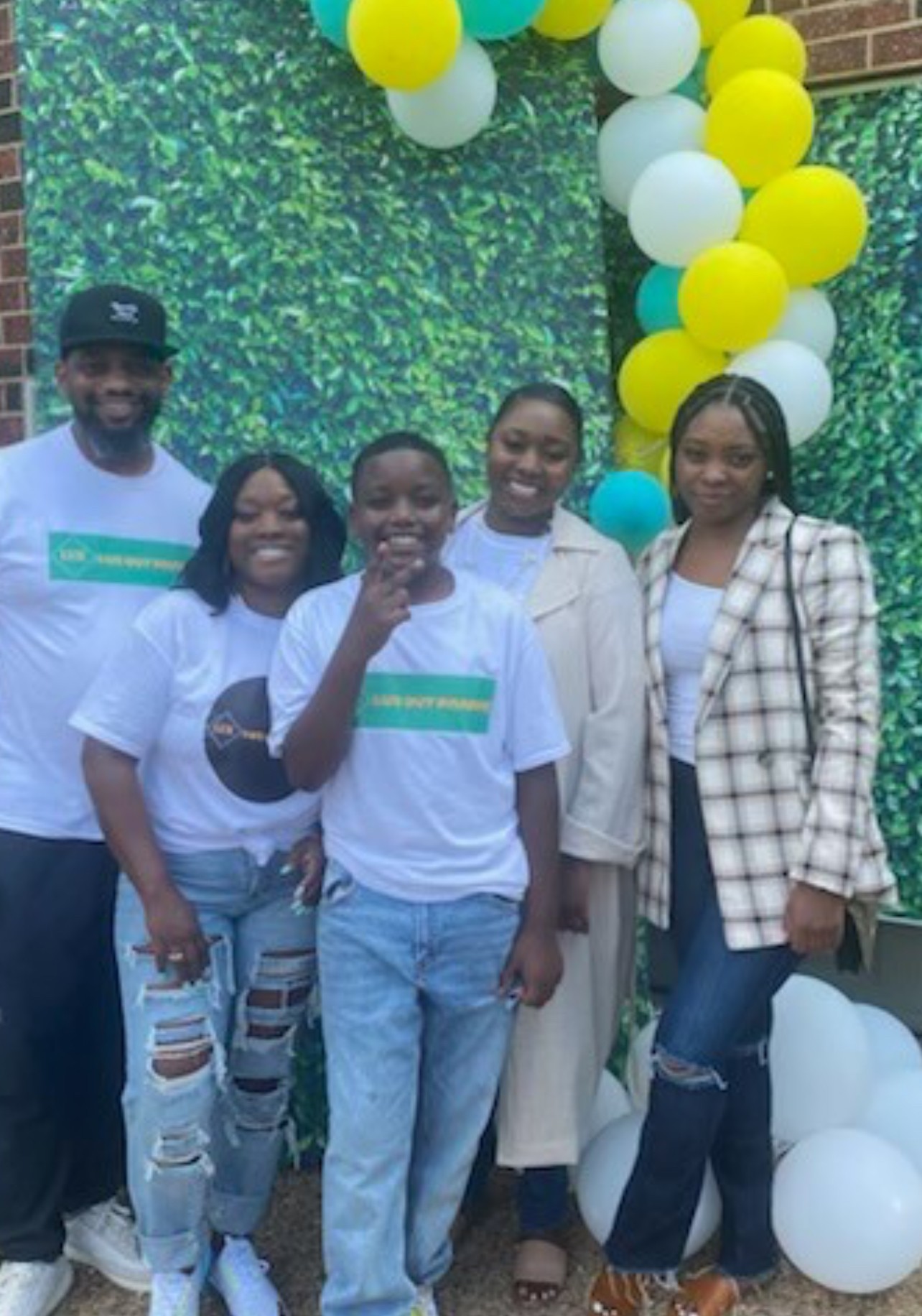 Group of five people smiling, standing in front of a green leafy backdrop with yellow and white balloons, outdoors.