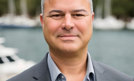 Man in a gray suit and light blue shirt outdoors with boats and water in the background.