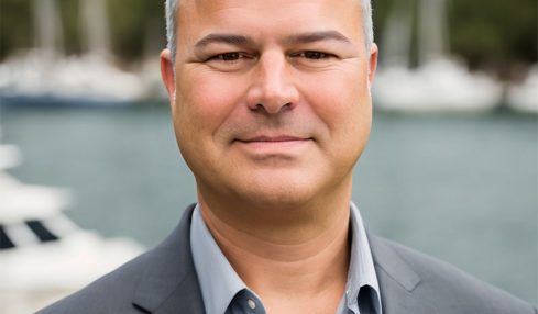 Man in a gray suit and light blue shirt outdoors with boats and water in the background.