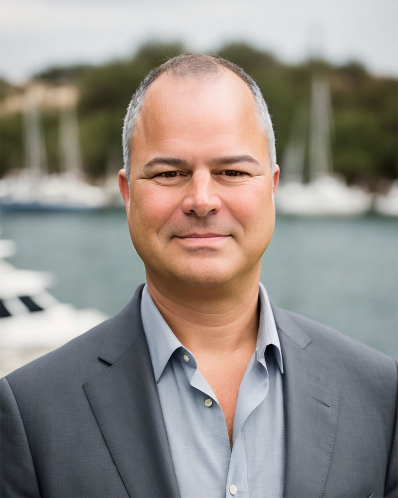 Man in a gray suit and light blue shirt outdoors with boats and water in the background.