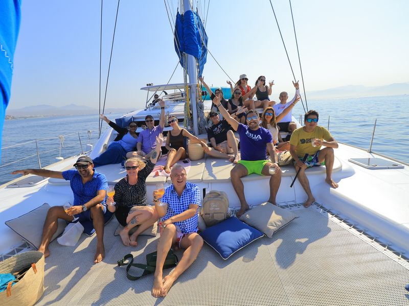 Group of people on a boat deck, some sitting and some standing, with a calm body of water in the background.