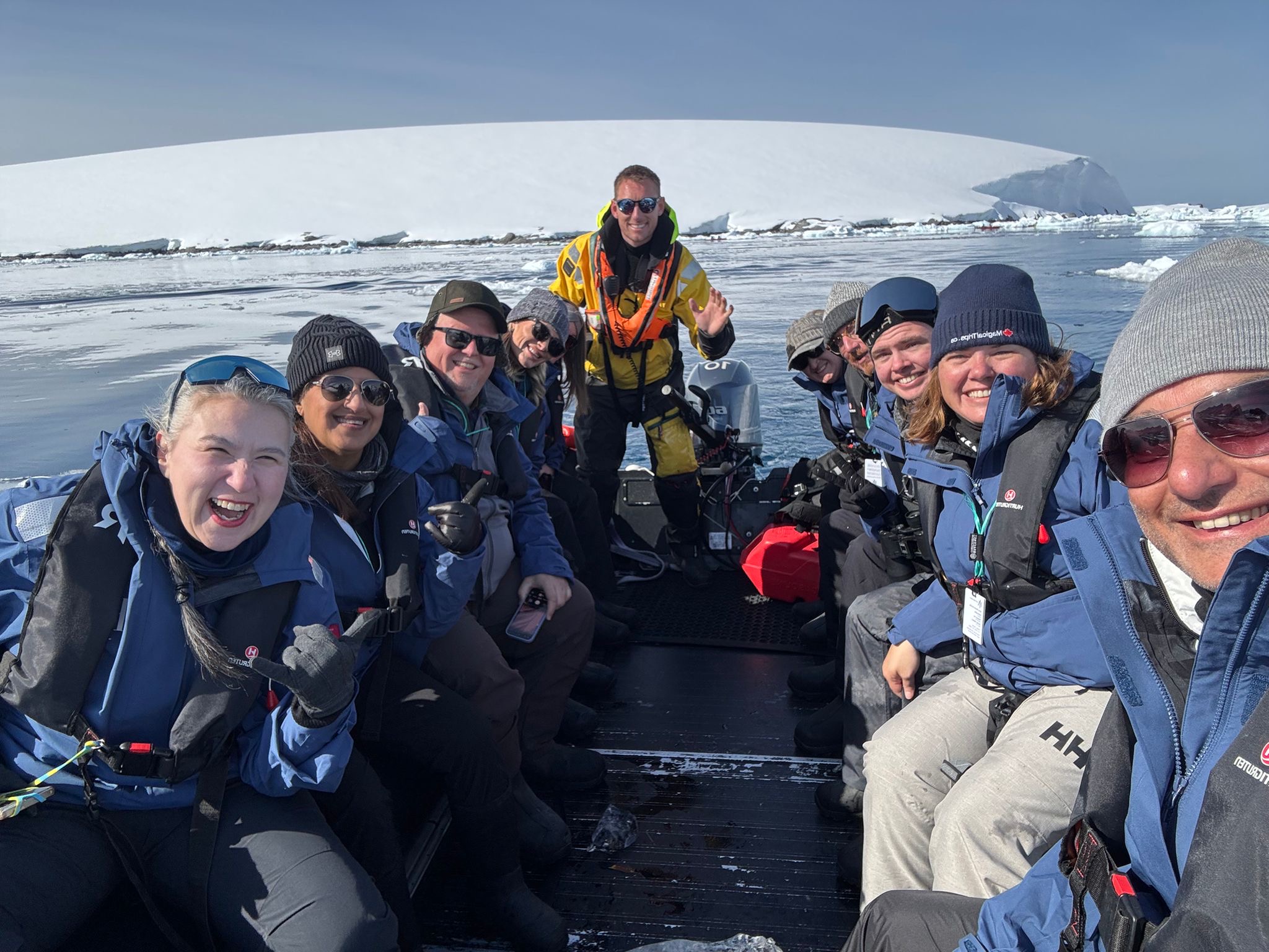 Group of people in outdoor gear sitting on a boat in icy water, with icebergs in the background, smiling at the camera.