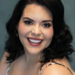 Smiling woman with dark wavy hair, makeup, and a sleeveless top, against a teal background.