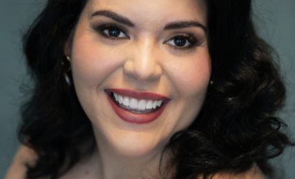 Smiling woman with dark wavy hair, makeup, and a sleeveless top, against a teal background.