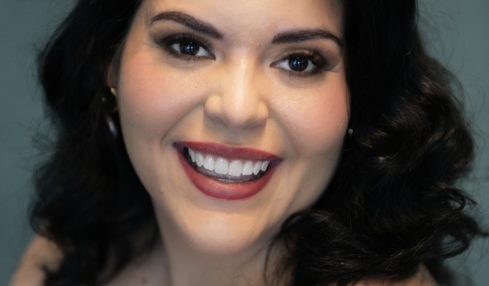 Smiling woman with dark wavy hair, makeup, and a sleeveless top, against a teal background.