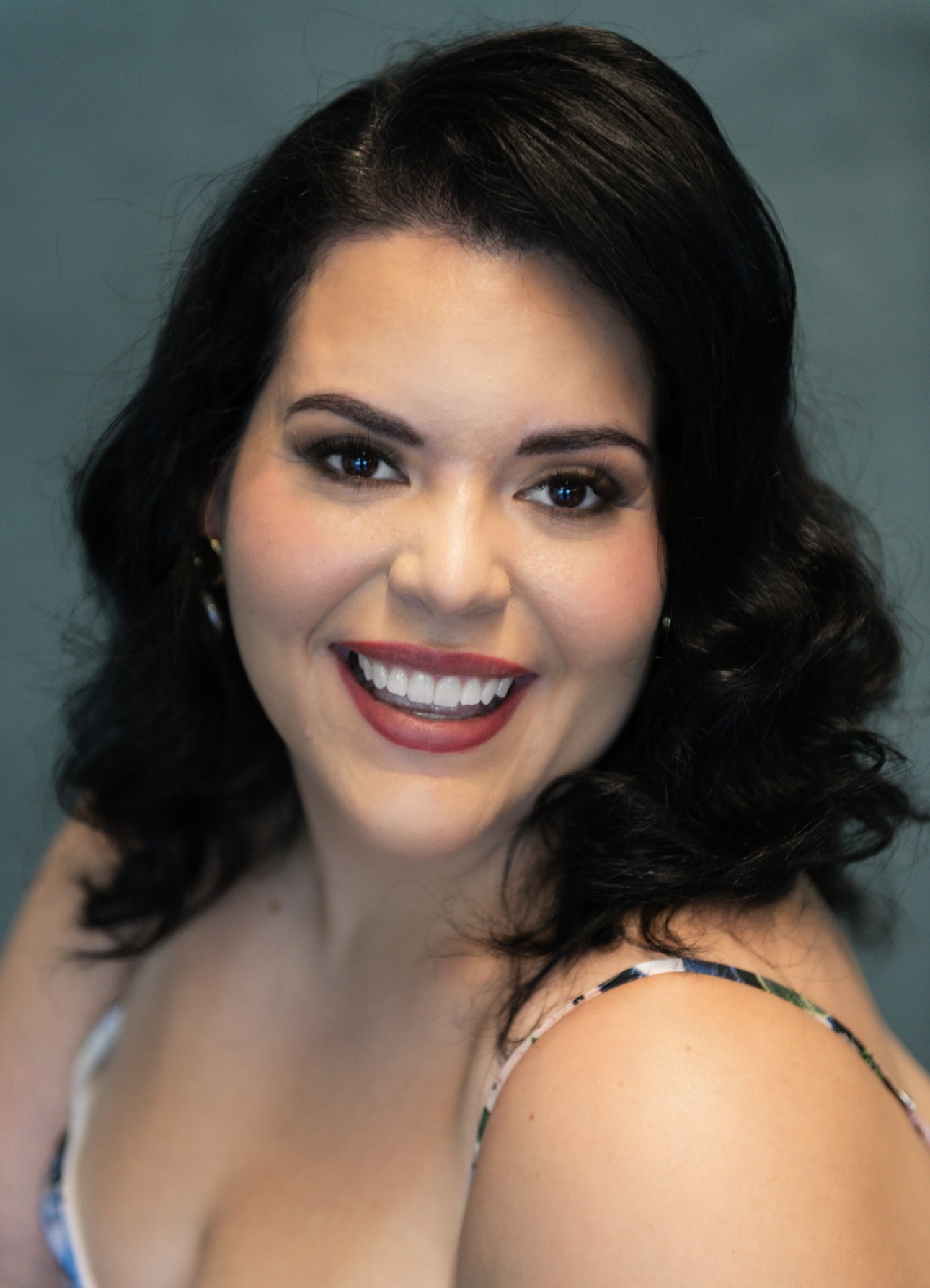 Smiling woman with dark wavy hair, makeup, and a sleeveless top, against a teal background.