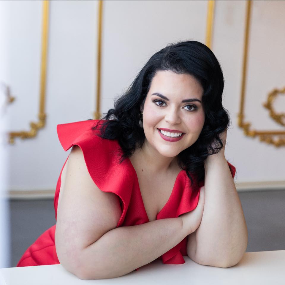 Woman with dark hair and red dress smiling, resting her head on her hand, in an elegant room.