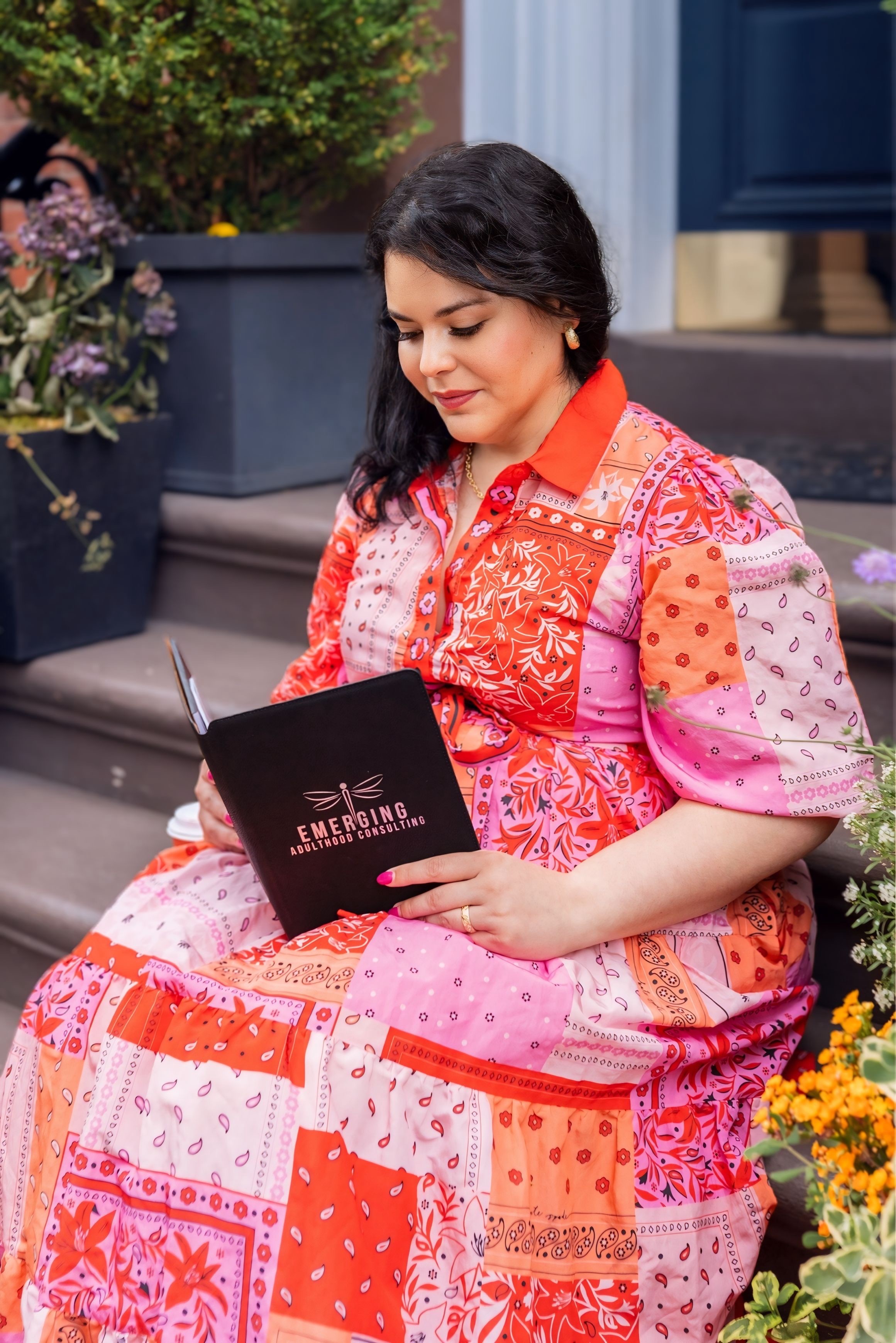 Woman sitting outdoors reading a book, wearing a colorful patchwork dress, with plants and flowers nearby.