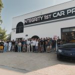 Group of people standing outside a building with a sign reading 'INTEGRITY 1ST CAR PROS' and a black sports car in front.