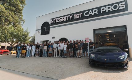 Group of people standing outside a building with a sign reading 'INTEGRITY 1ST CAR PROS' and a black sports car in front.