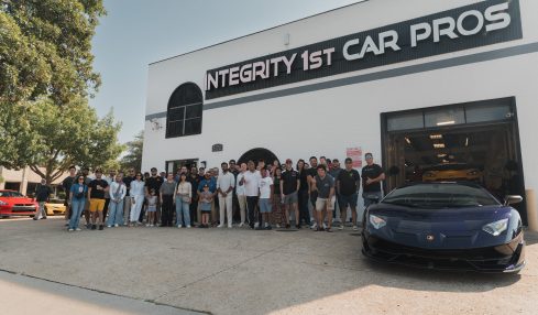 Group of people standing outside a building with a sign reading 'INTEGRITY 1ST CAR PROS' and a black sports car in front.
