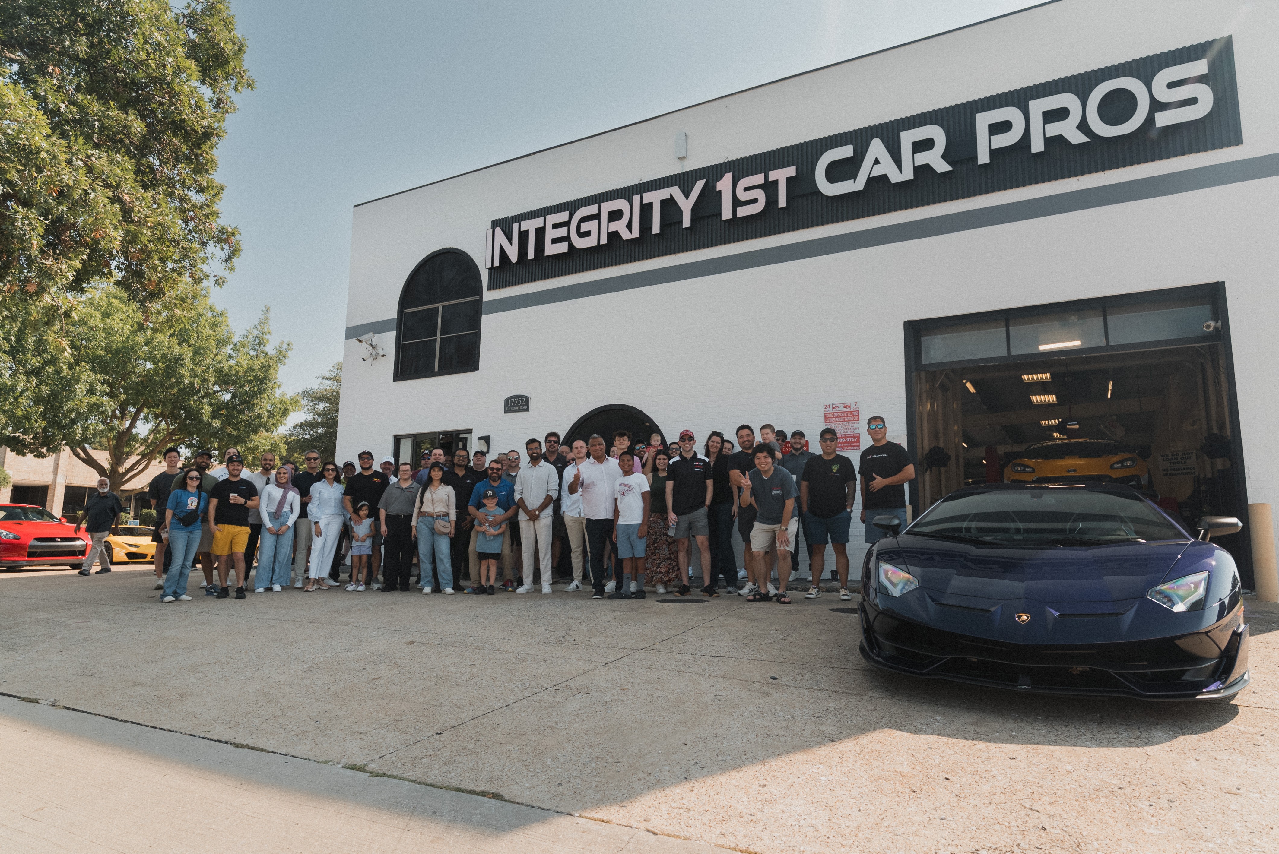 Group of people standing outside a building with a sign reading 'INTEGRITY 1ST CAR PROS' and a black sports car in front.