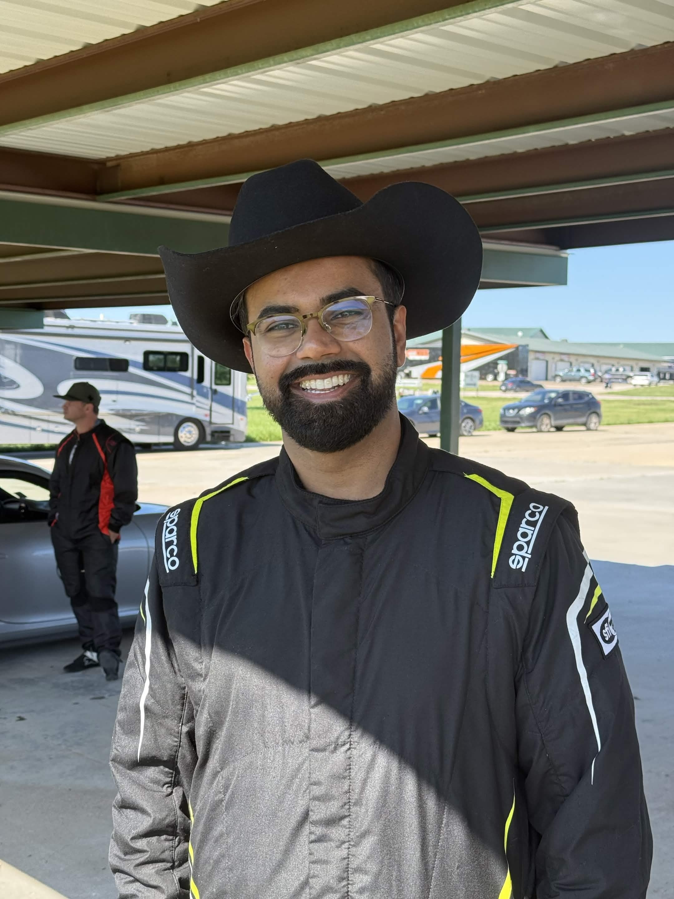 Man with glasses and a beard wearing a black cowboy hat and racing suit, smiling outdoors under a shelter.