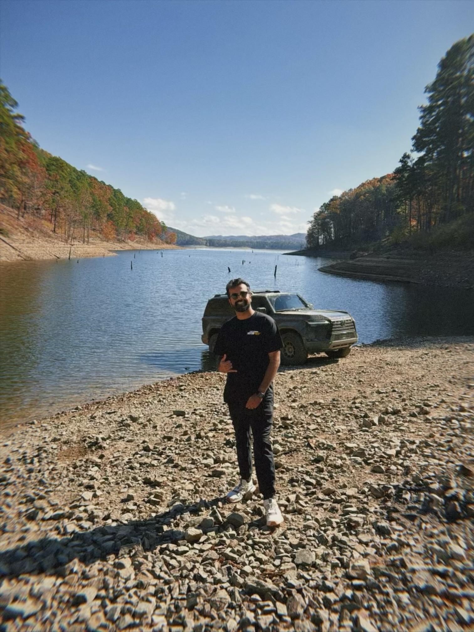 Person standing on rocky shore near a car by a lake with trees and blue sky in background.