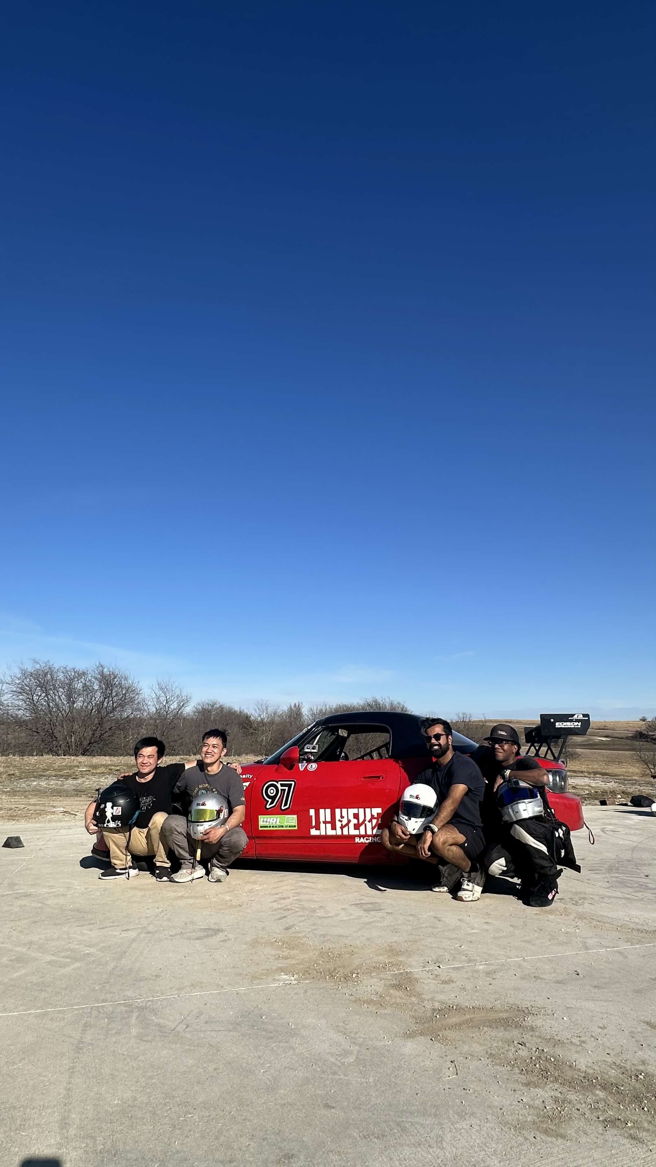 Three people kneeling beside a red vehicle and a motorcycle outdoors under a clear blue sky.