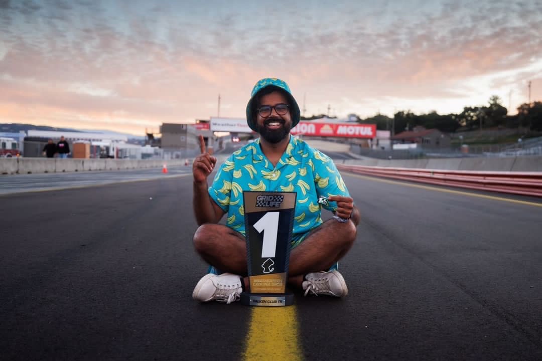 Person sitting on a race track holding a sign with the number one, smiling, with a sunset sky in the background.