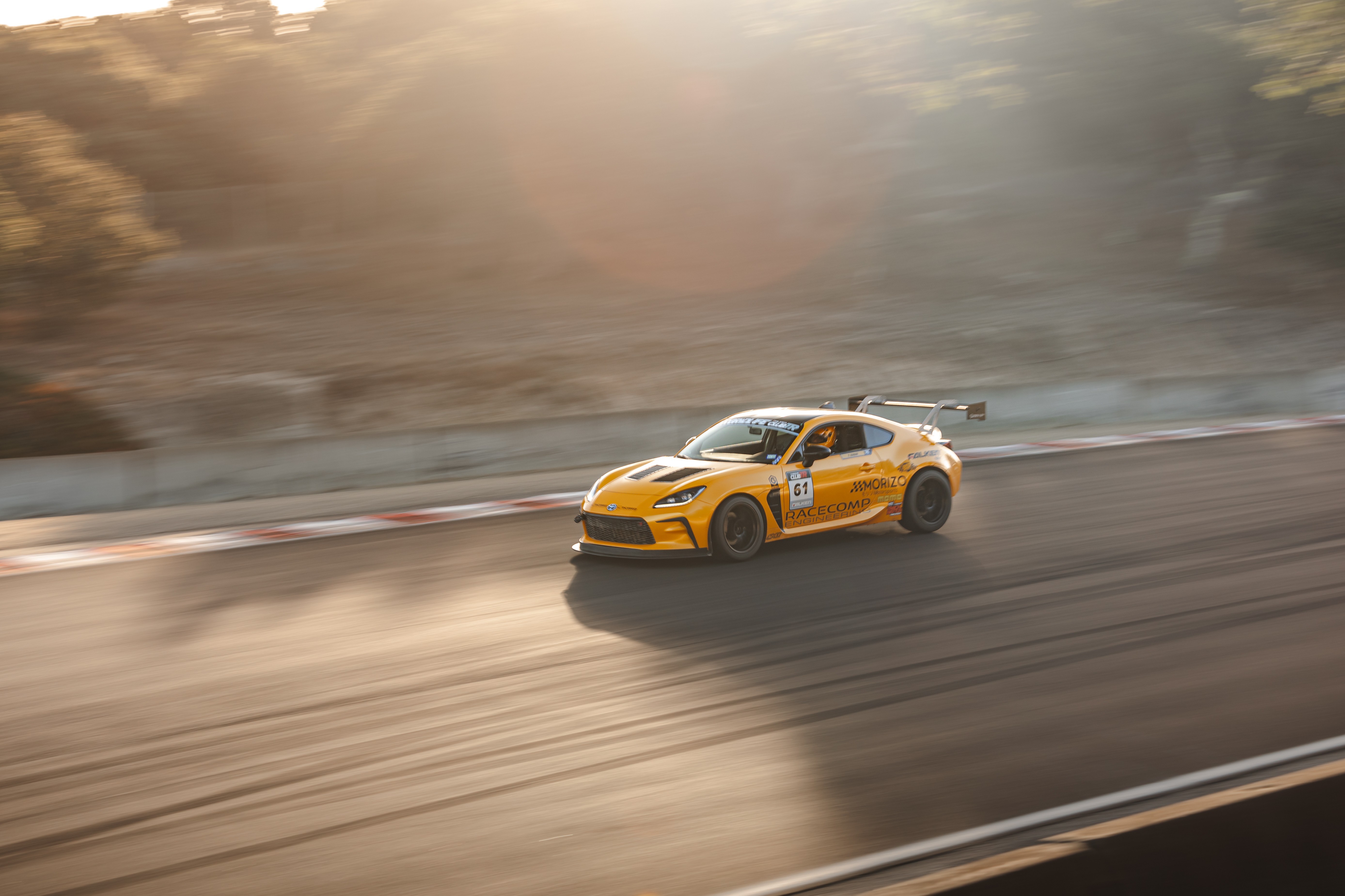 Yellow race car on a track with motion blur, sunlight shining from above, and trees in the background.