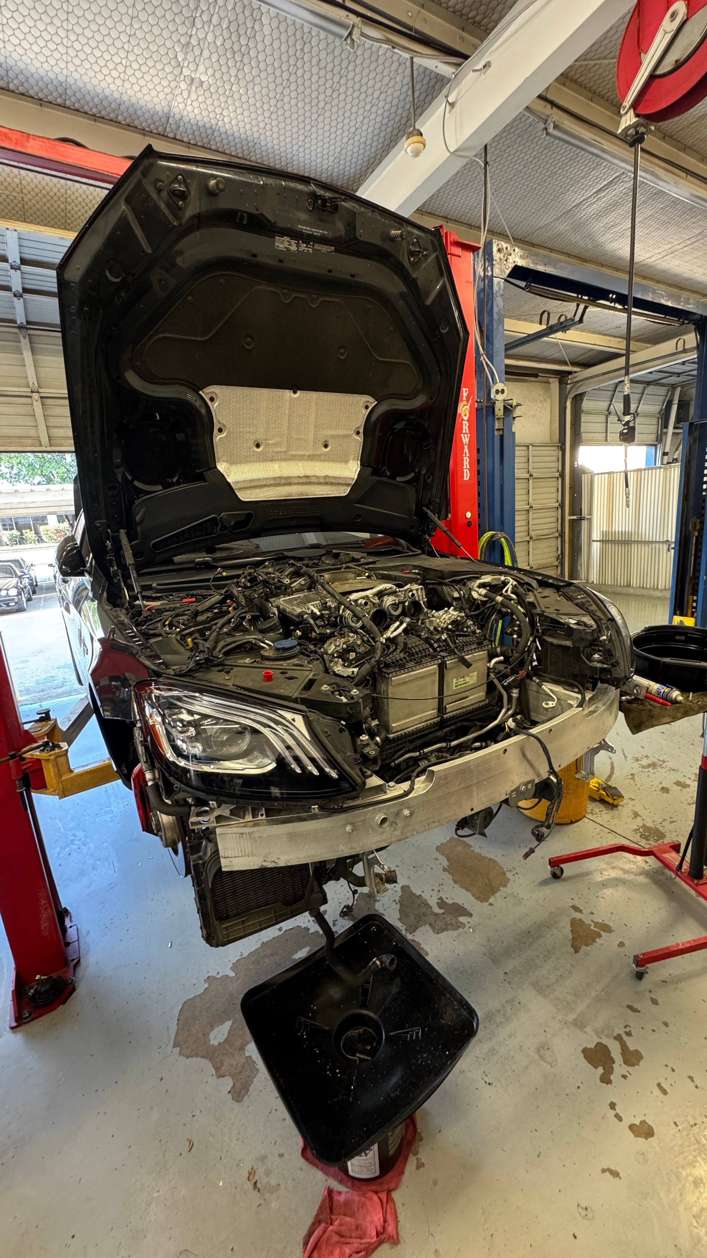 Car with open hood in garage, engine visible, tools nearby, floor with oil stain, garage door in background.