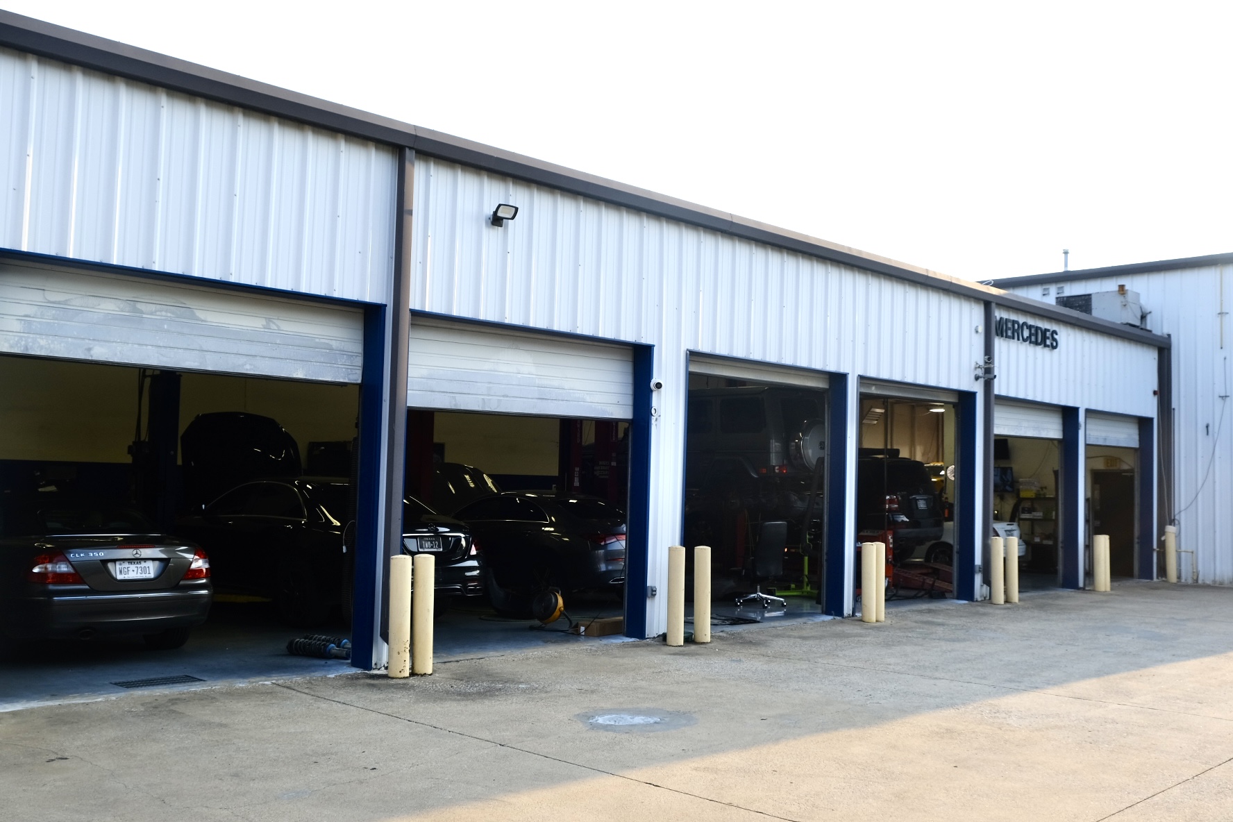 Automotive repair shop with multiple garage bays, cars parked inside, and yellow safety bollards outside, under a metal building.