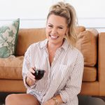 Smiling woman with blonde hair in a ponytail, wearing a striped shirt, holding a glass of red wine, sitting on a sofa.