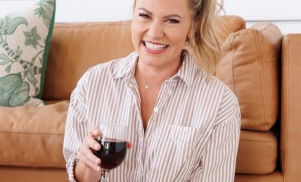 Smiling woman with blonde hair in a ponytail, wearing a striped shirt, holding a glass of red wine, sitting on a sofa.