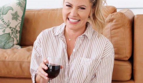 Smiling woman with blonde hair in a ponytail, wearing a striped shirt, holding a glass of red wine, sitting on a sofa.