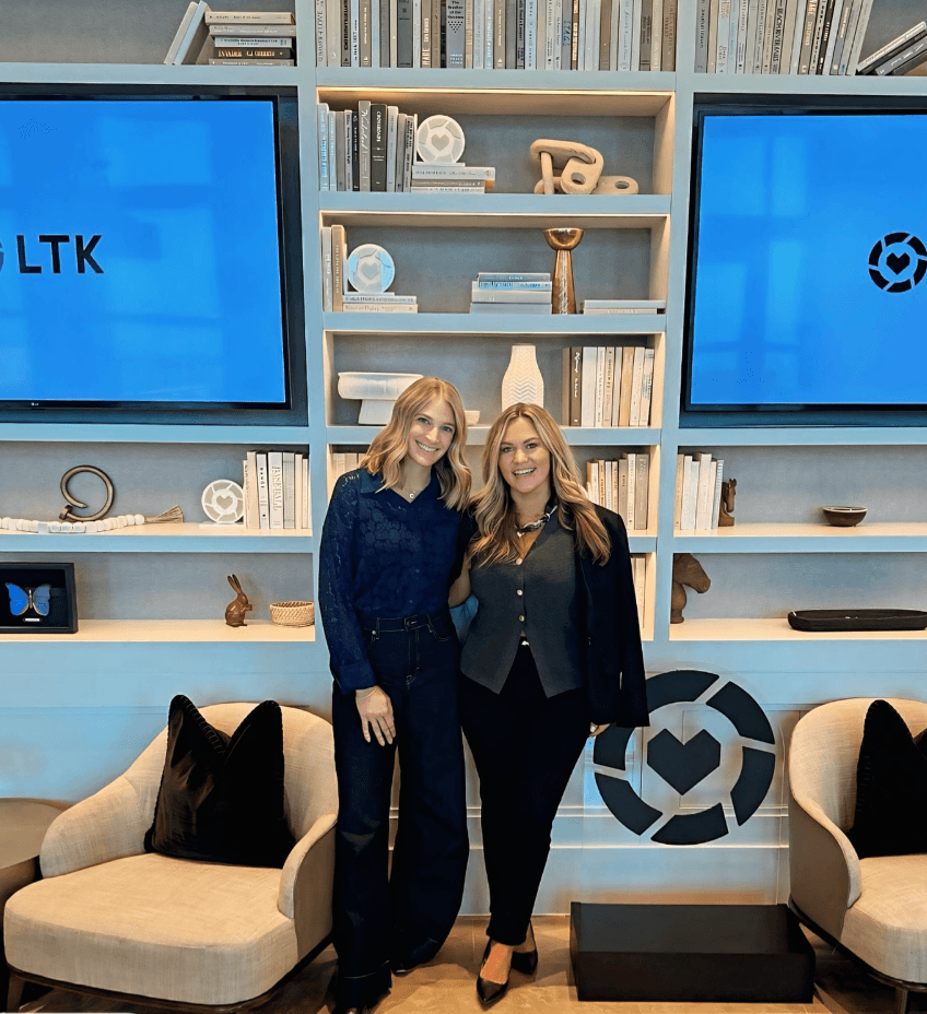 Two women standing in front of a blue bookshelf with decorative items and two screens, smiling at the camera.