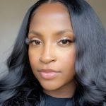 Close-up portrait of a woman with long, wavy black hair and a nose piercing, looking directly at the camera.