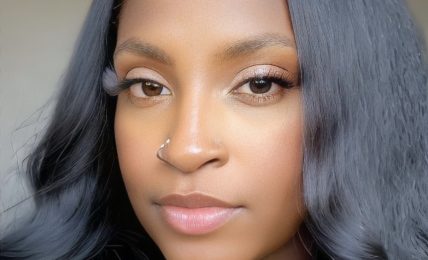 Close-up portrait of a woman with long, wavy black hair and a nose piercing, looking directly at the camera.