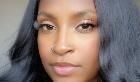 Close-up portrait of a woman with long, wavy black hair and a nose piercing, looking directly at the camera.