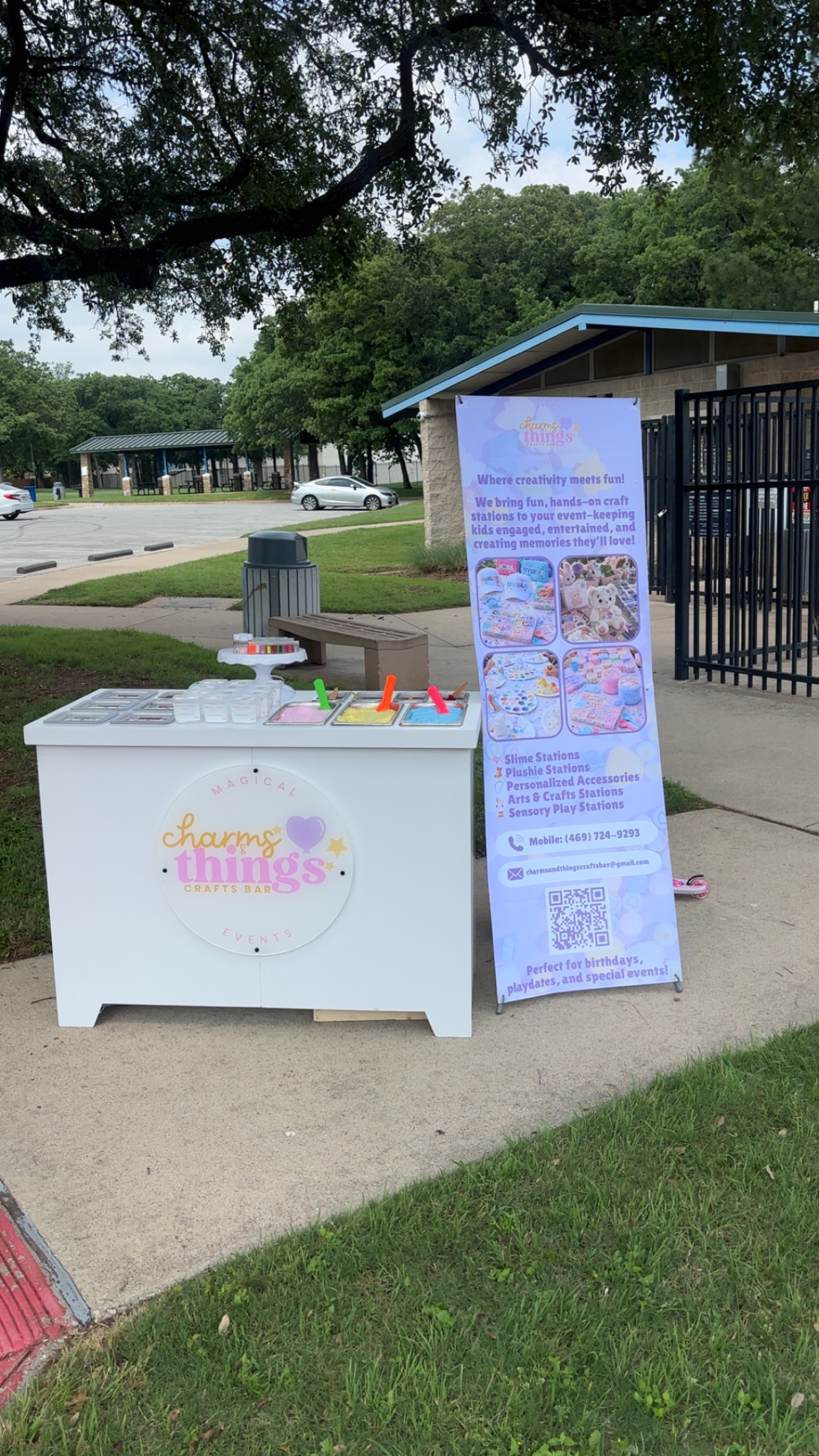 Table with colorful items and a tall informational banner outside near a building and parking lot.