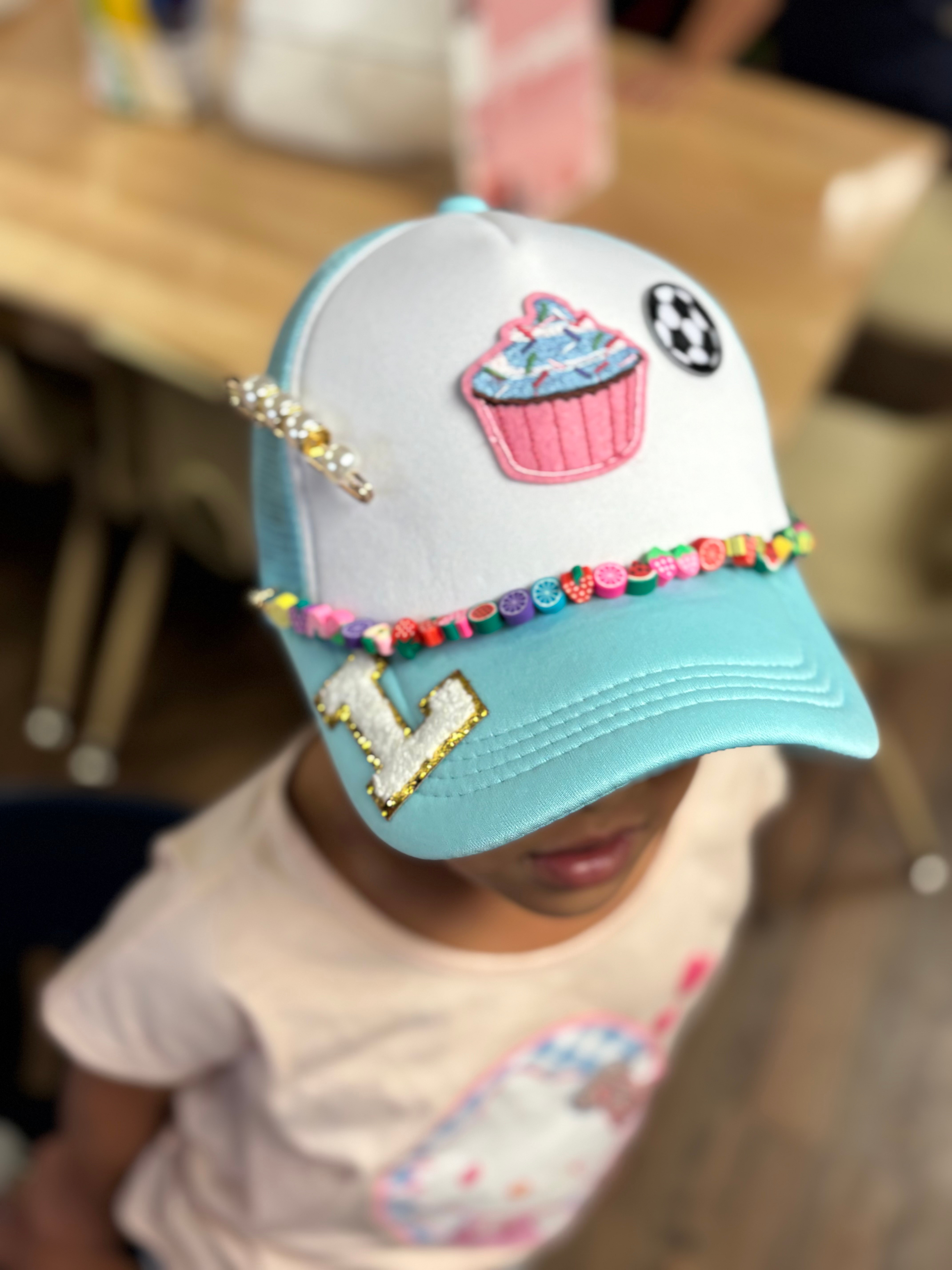 Child wearing a white and blue cap with cupcake and star patches, decorated with beads and charms, in a room.